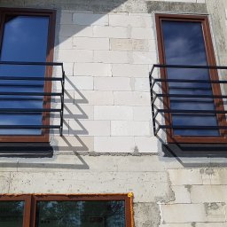 ArtisOmnia - Dwa okna z brązowymi ramami zabezpieczone czarnymi, prostymi balustradami metalowymi, zamontowane na surowej ścianie z białych bloczków.