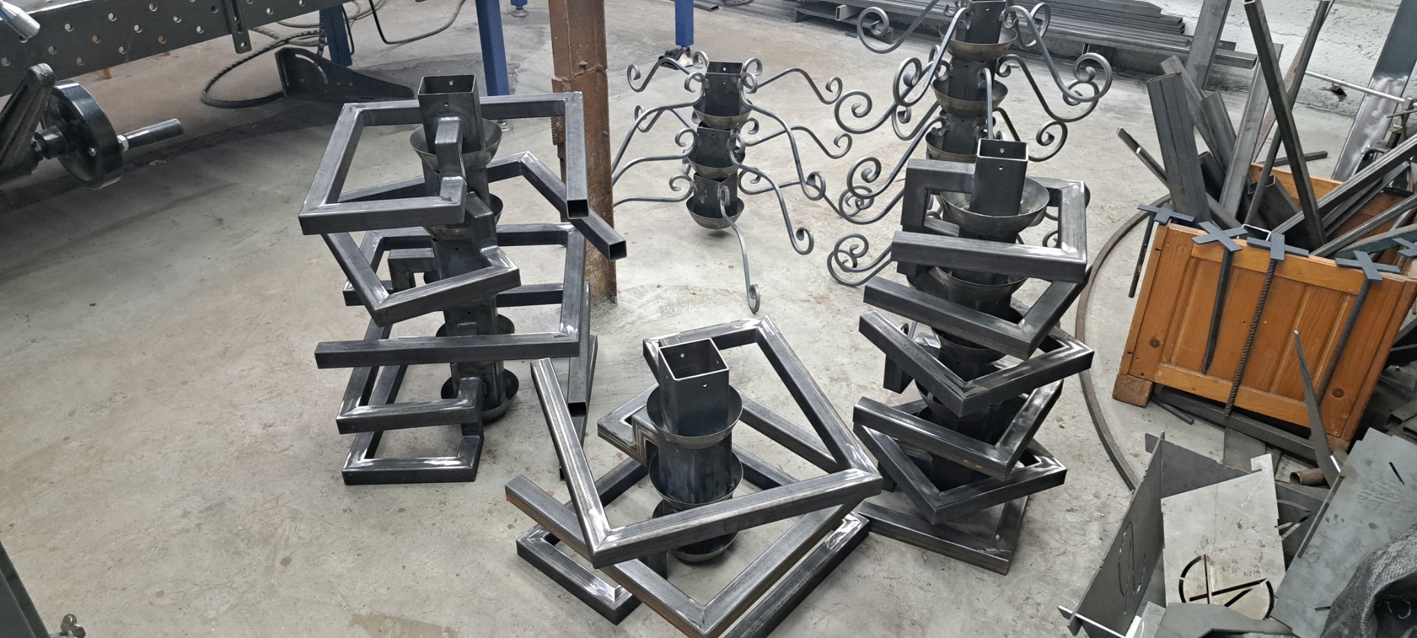 Nieskończone metalowe konstrukcje spawane z profili kwadratowych, tworzące geometryczne formy przypominające podstawy lamp lub kwietników, widoczne w warsztacie z narzędziami i skrzynią...