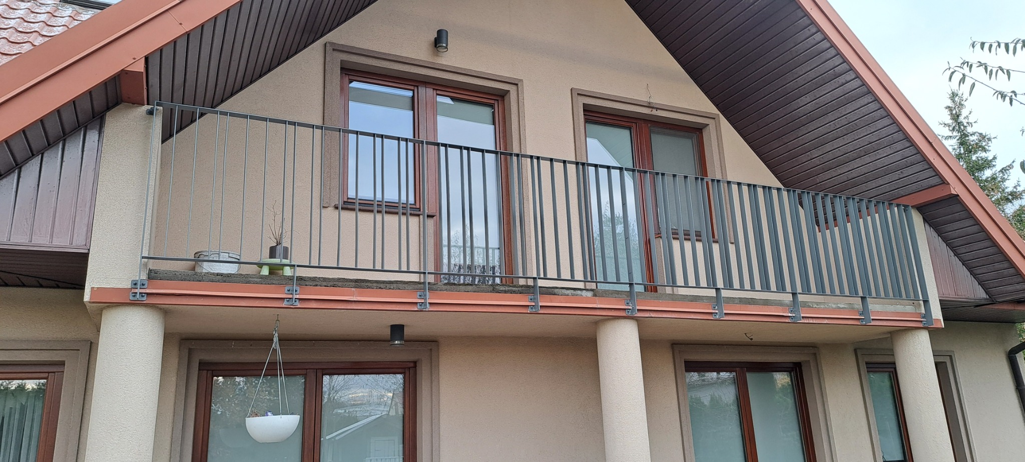 Balkon z metalową balustradą w kolorze grafitowym, mocowaną na brązowych elementach konstrukcyjnych. Na balkonie doniczki z roślinami, widok na elewację domu z drewnianymi oknami i brązowym dachem.