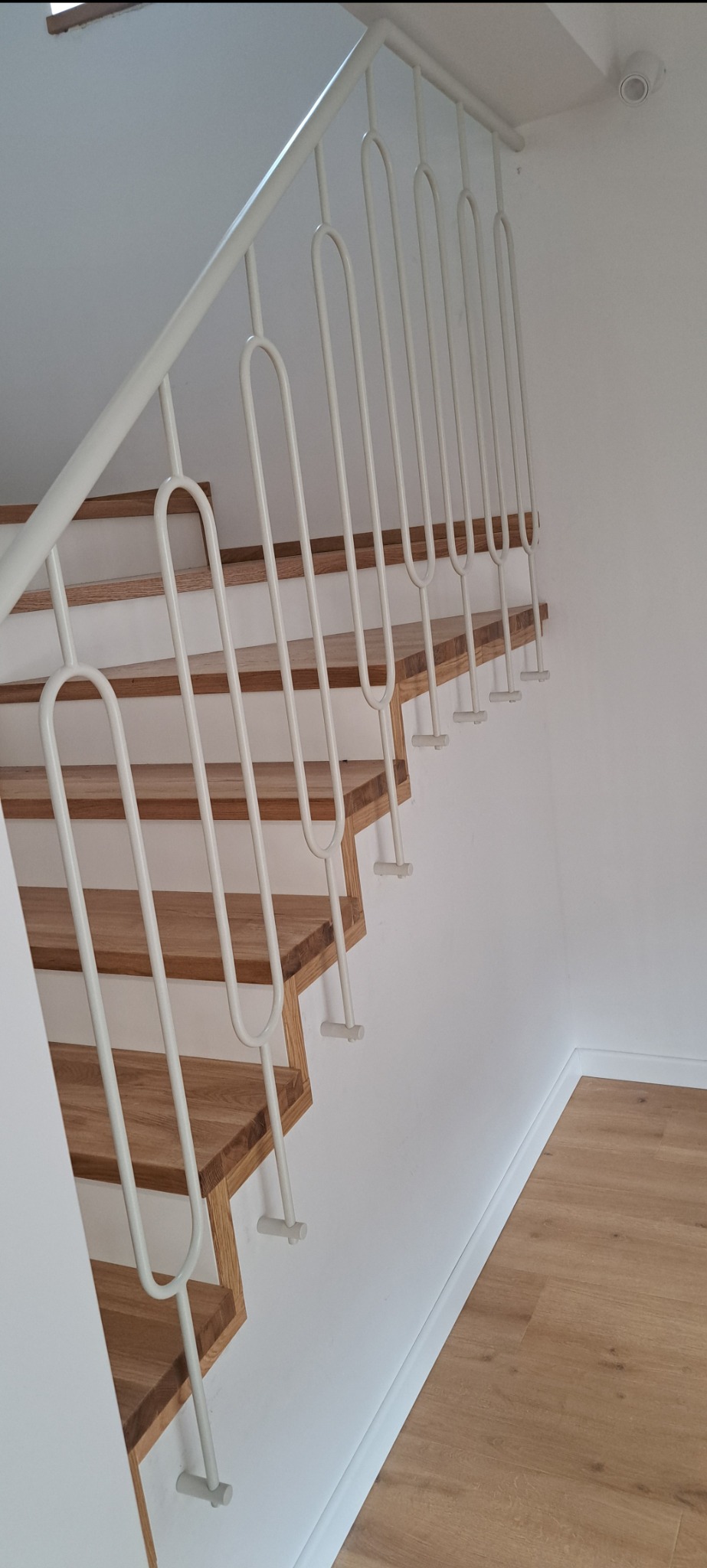 Schody drewniane z białą, metalową balustradą o minimalistycznym designie, z charakterystycznymi zaokrąglonymi elementami pionowymi.