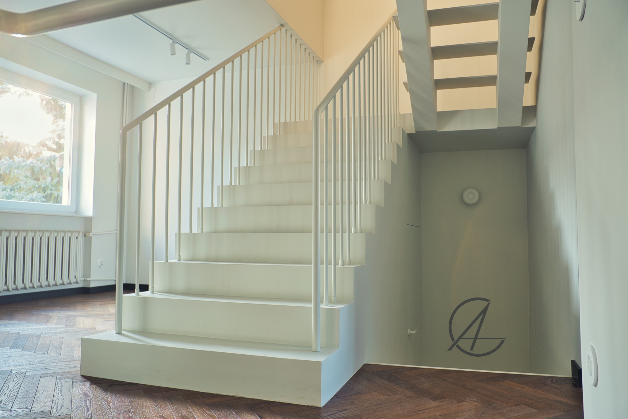Nowoczesne, białe schody z metalową balustradą w jasnym wnętrzu z parkietem w jodełkę. Widoczny fragment okna z grzejnikiem i logo firmy na ścianie pod schodami.