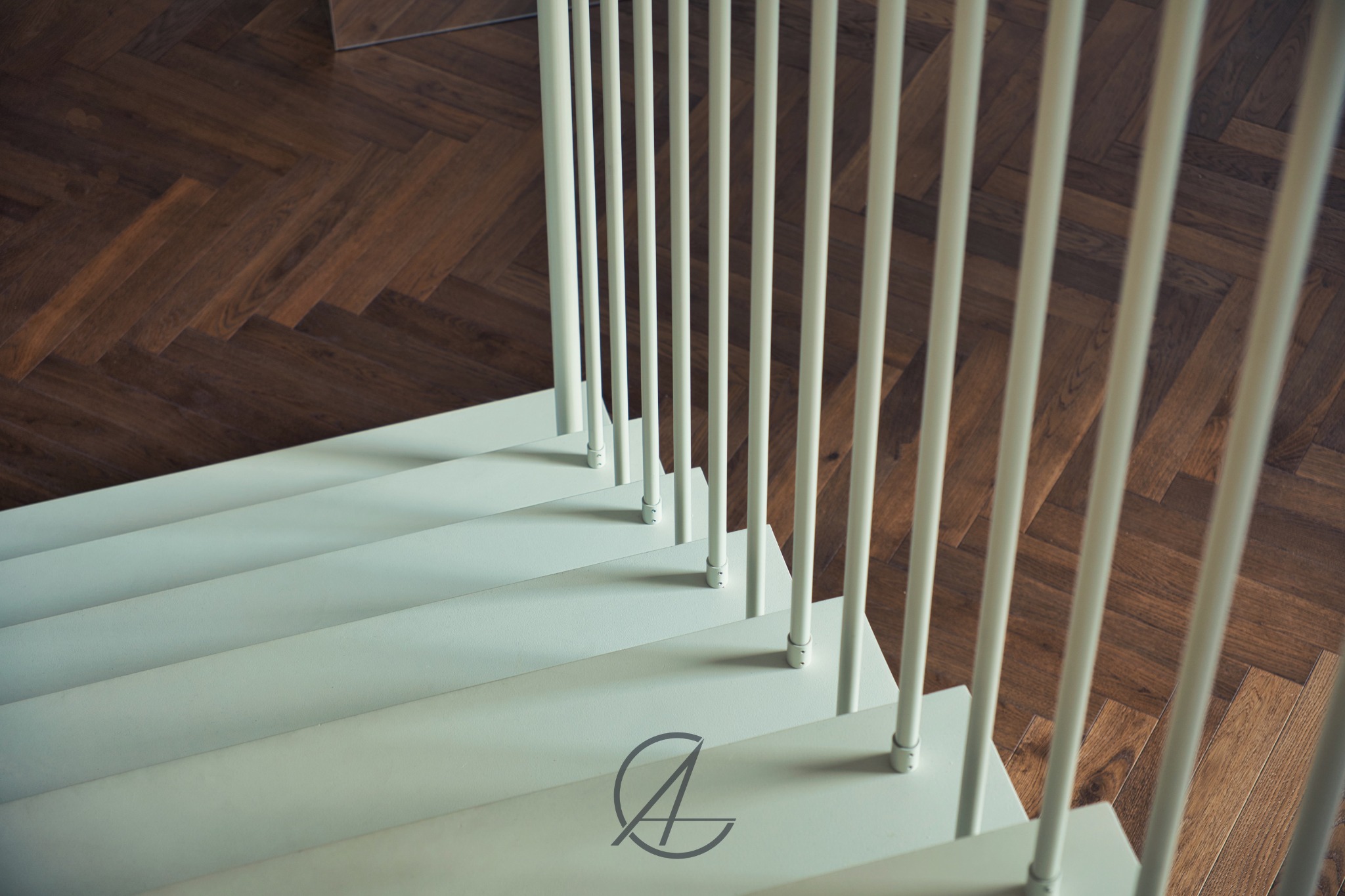 Białe schody z metalową balustradą z okrągłych prętów, widok z góry na parkiet ułożony w jodełkę, na pierwszym planie logo firmy.