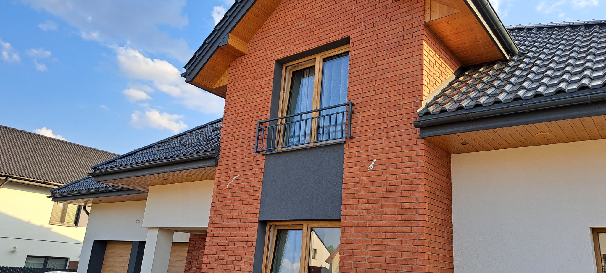 Nowoczesny dom z cegły klinkierowej z czarną balustradą balkonową i oknami w drewnianych ramach, widok z dołu na tle błękitnego nieba z chmurami.