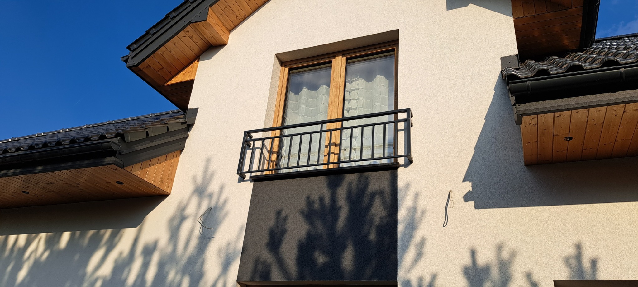 Czarna, prosta balustrada balkonowa z metalu, zamontowana przy oknie na elewacji budynku o jasnej fasadzie i drewnianych elementach dachu.