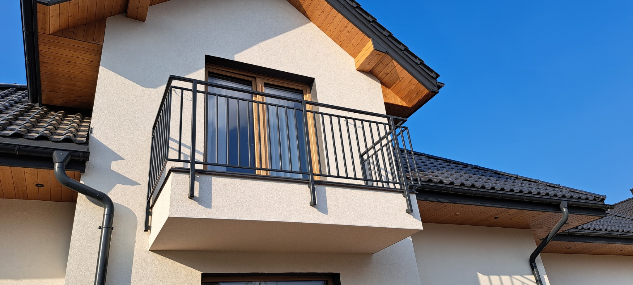 Balkon z czarną, metalową balustradą o minimalistycznym designie, zamontowany przy oknie na elewacji nowoczesnego domu z drewnianą podbitką dachową i czarną rynną, sfotografowany w słoneczny dzień.