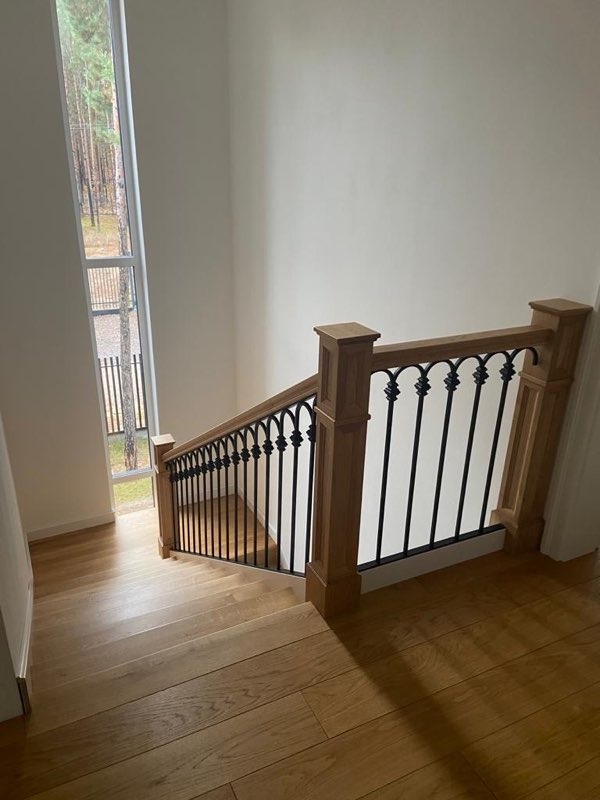 Schody drewniane z czarną, kutą balustradą, widok z góry na dół, naturalne światło z wysokiego okna, widok na las w tle.