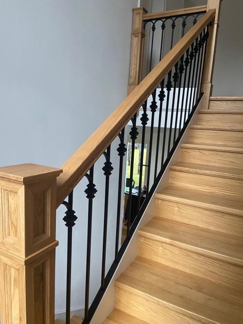 Drewniane schody z czarną, kutą balustradą z ozdobnymi elementami, widok z dołu, jasne wnętrze.