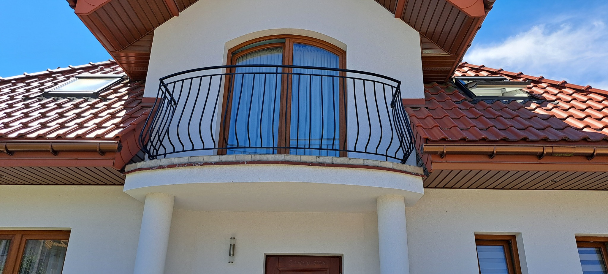 Czarna, ozdobna balustrada balkonowa z pionowymi prętami na białym budynku z brązowym dachem i oknami dachowymi.