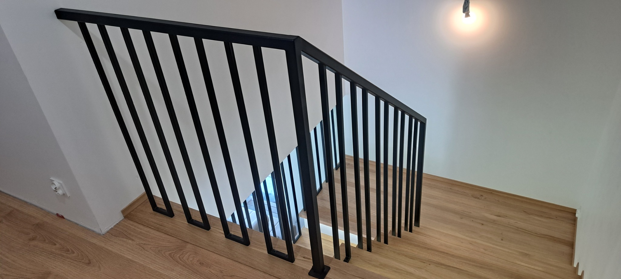 Nowoczesna, czarna balustrada schodowa z pionowymi szczeblami w minimalistycznym wnętrzu z drewnianymi schodami i białymi ścianami, oświetlona punktowym światłem.