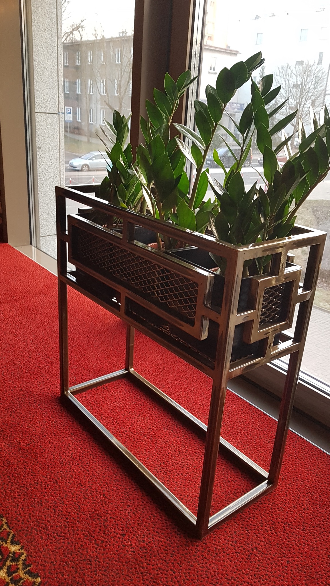 Metalowa konstrukcja kwietnika w stylu art deco, z roślinami zamiokulkas, na tle okna i czerwonej wykładziny.