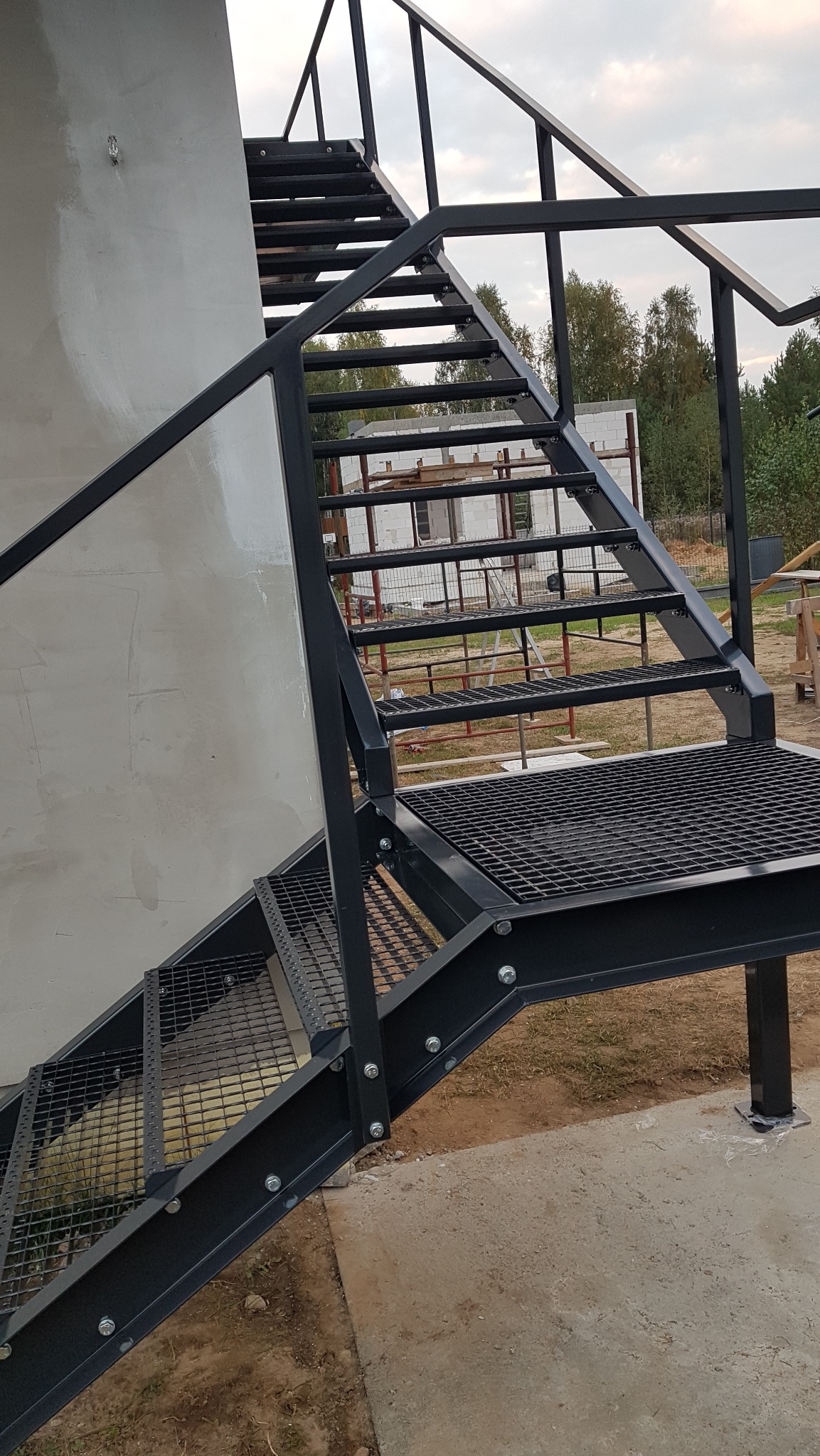 Zewnętrzne schody metalowe z platformą, pomalowane na czarno, z kratowymi stopniami, przymocowane do szarej ściany budynku w trakcie budowy.