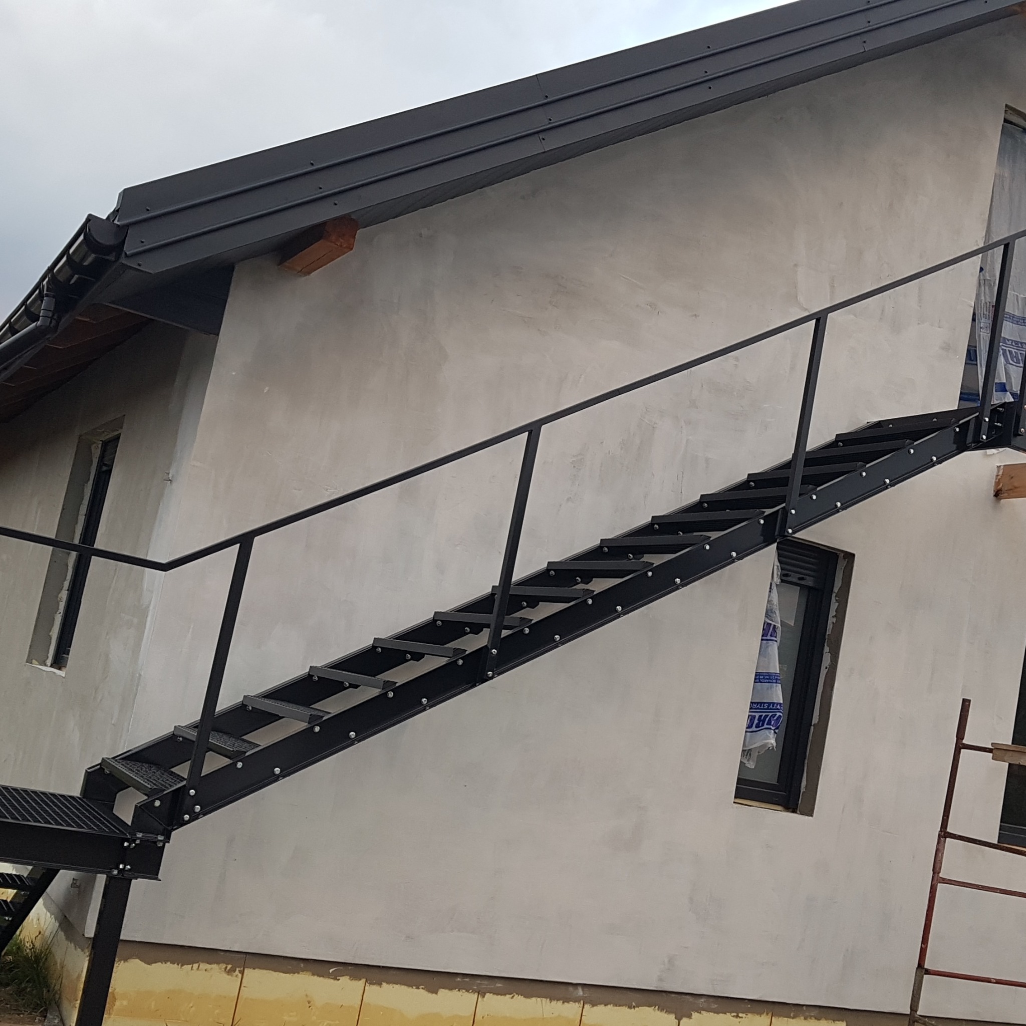 Zewnętrzne, czarne schody metalowe z poręczą, prowadzące do budynku z szarą elewacją, widoczne okna zabezpieczone folią i drabina oparta o ścianę.