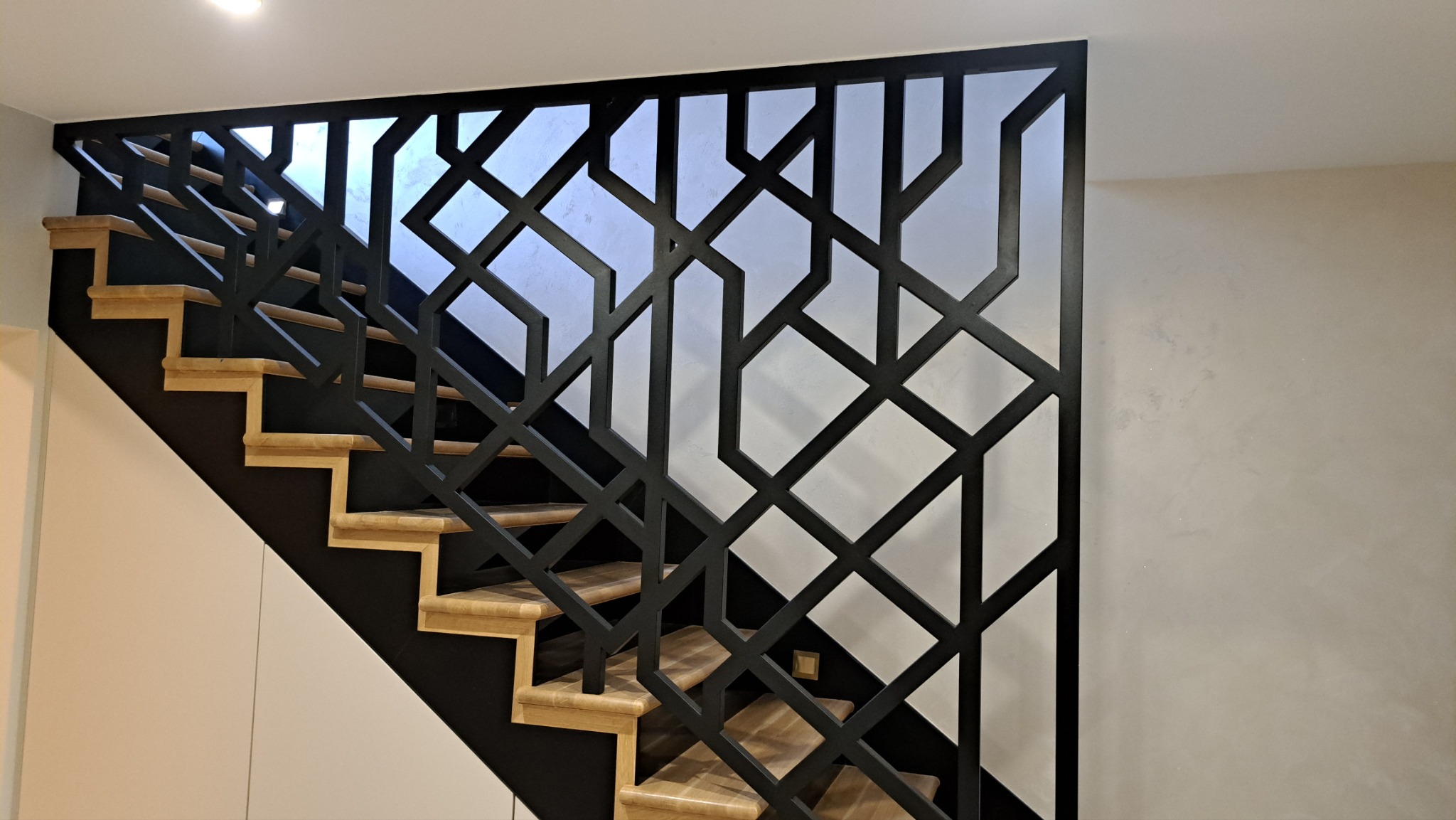 Nowoczesne schody z drewnianymi stopniami i geometryczną, czarną balustradą metalową, widok z dołu na jasne wnętrze.