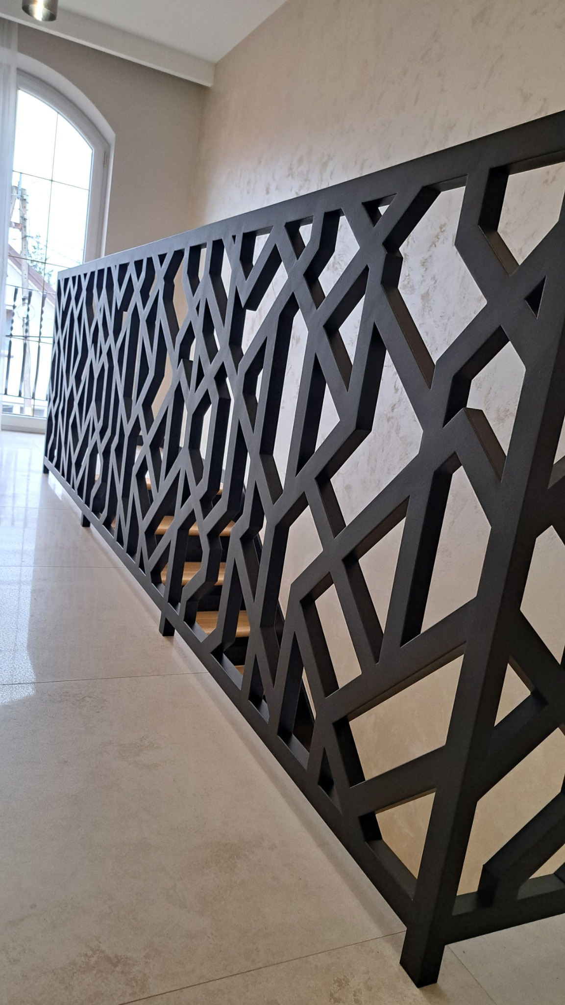 Czarna, metalowa balustrada schodowa o geometrycznym wzorze, widziana z dołu pod kątem, z widocznymi stopniami schodów i oknem w tle.