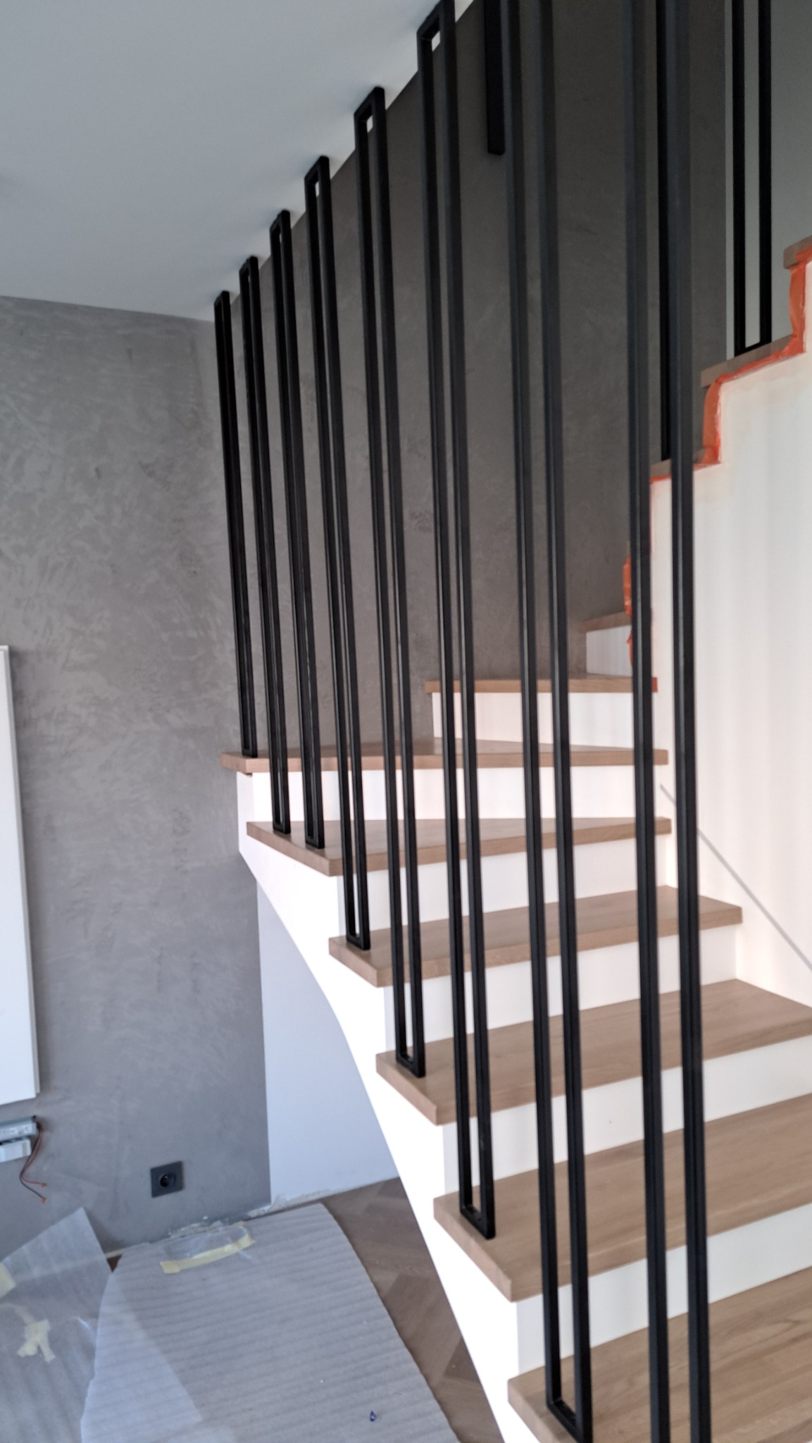 Nowoczesne schody z drewnianymi stopniami i minimalistyczną, czarną balustradą z pionowych prętów na tle szarej ściany o fakturze betonu, zabezpieczone folią podłogi.