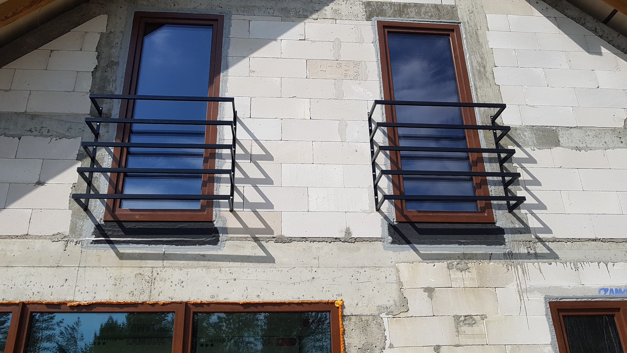 Dwa okna z brązowymi ramami zabezpieczone czarnymi, prostymi balustradami metalowymi, zamontowane na surowej ścianie z białych bloczków.