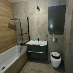 Nowoczesna łazienka z szarymi płytkami imitującymi kamień, podświetlanym sufitem LED, kabiną prysznicową nad wanną z czarnymi elementami, szarą szafką pod umywalką i podwieszaną szafką nad toaletą...