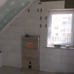 Łazienka w trakcie wykańczania: częściowo położone jasne płytki ścienne z krzyżykami dystansowymi, zabudowa stelaża WC z okładziną drewnopodobną, okno z widokiem na dom.