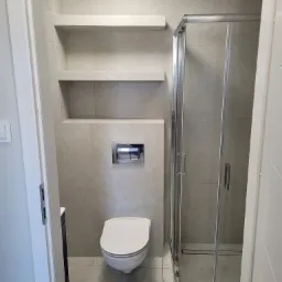 Nowoczesna łazienka w beżowych płytkach z podwieszaną toaletą, kabiną prysznicową ze srebrnymi profilami i półkami ściennymi.