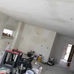 Wnętrze w trakcie remontu: ściany po gładziowaniu, widoczne narzędzia, wiadra z zaprawą, worki z materiałami budowlanymi i drabina, przygotowane do dalszych prac wykończeniowych.