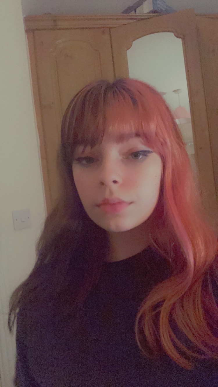 Portret młodej kobiety z brązowo-różowymi włosami na tle jasnej ściany i drewnianej szafy z lustrem.