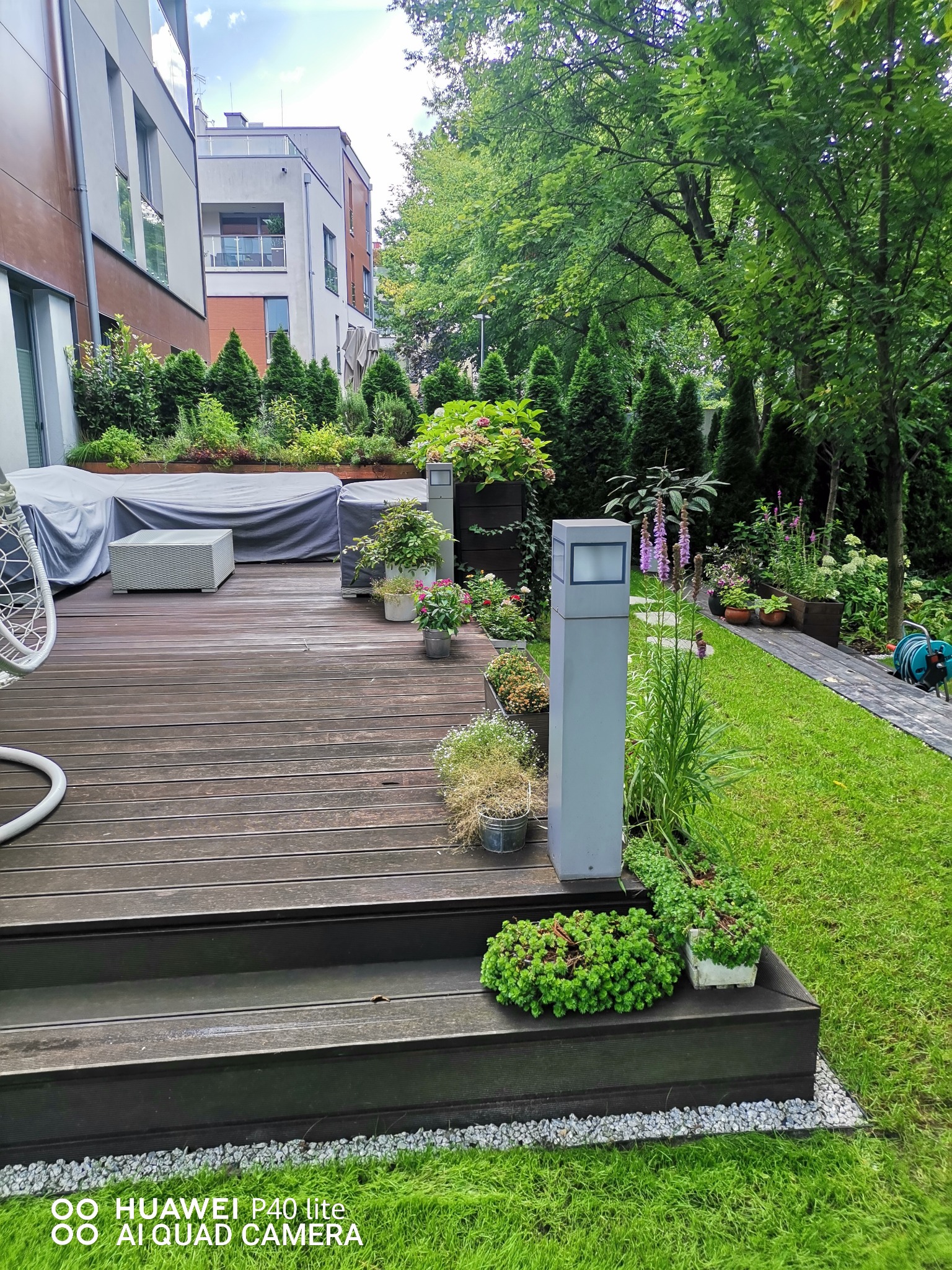 Ujęcie z poziomu gruntu na taras z drewnianą podłogą i schodami, otoczony zielenią, z meblami ogrodowymi pod pokrowcem i donicami z kwiatami, oświetlony nowoczesną lampą ogrodową.