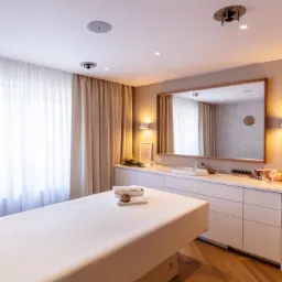 Jasne wnętrze gabinetu z łóżkiem do masażu, ręcznikiem z napisem 'slow', umywalką, lustrem w drewnianej ramie i drewnianą podłogą.