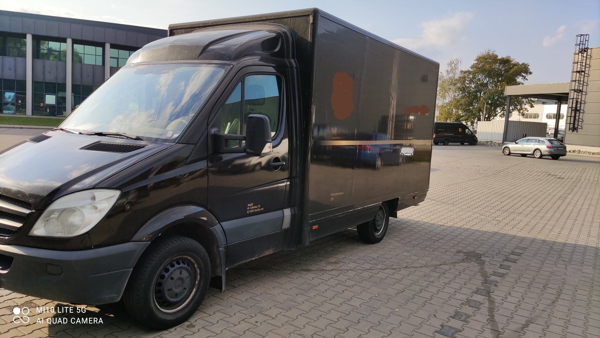 Czarny bus dostawczy UPS zaparkowany na kostce brukowej przed budynkiem biurowym w słoneczny dzień. Widoczny napis 'Podwykonawca UPS' oraz adres firmy.