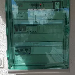 Rozdzielnica elektryczna z zieloną, przezroczystą obudową, zamontowana na białej, teksturowanej ścianie zewnętrznej, z widocznymi bezpiecznikami i opisami sektorów.