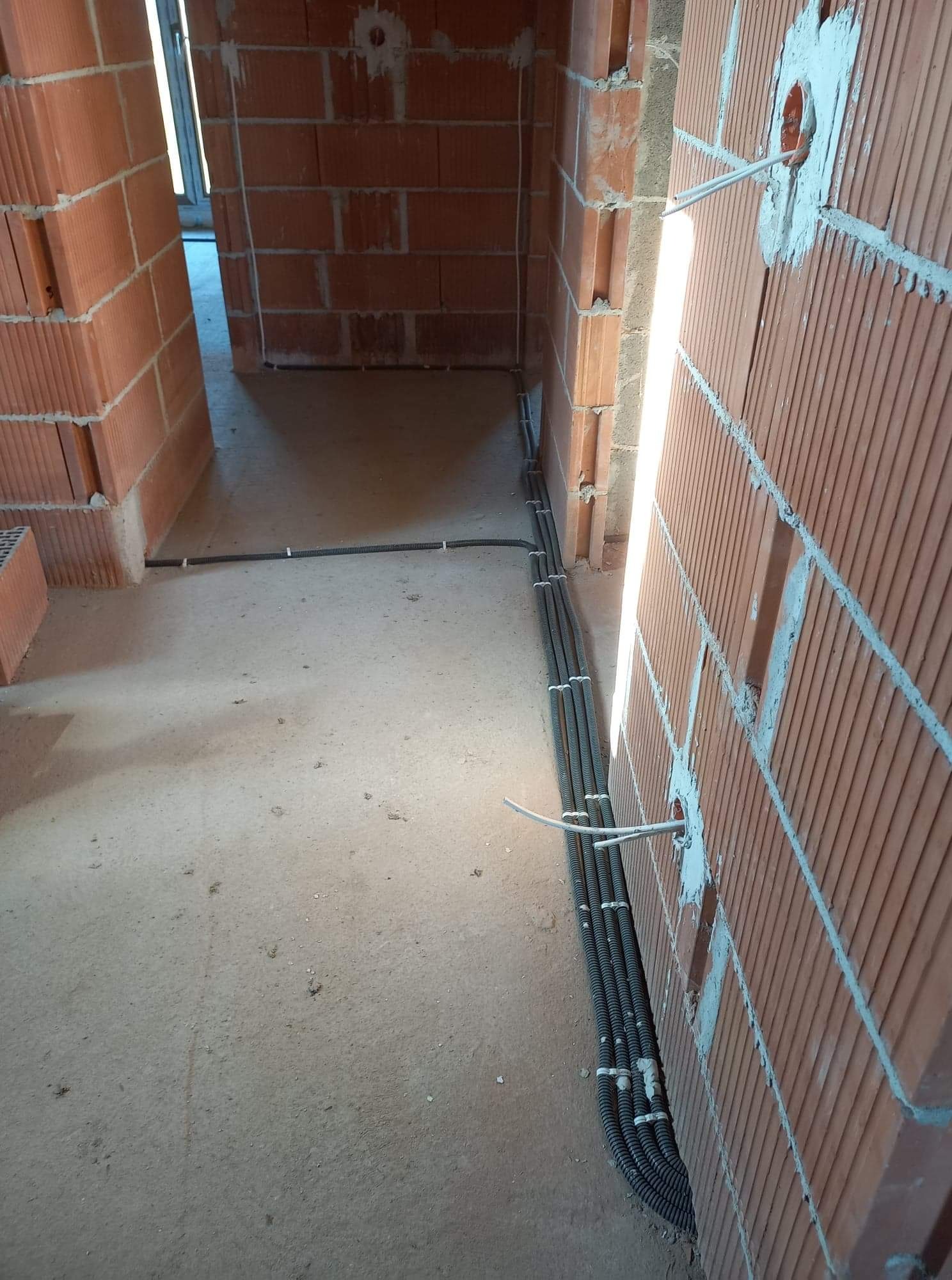 Wnętrze w trakcie budowy z widocznymi przewodami elektrycznymi w peszlach, prowadzonymi wzdłuż ściany z cegły, z wyprowadzonymi przewodami do gniazdek.
