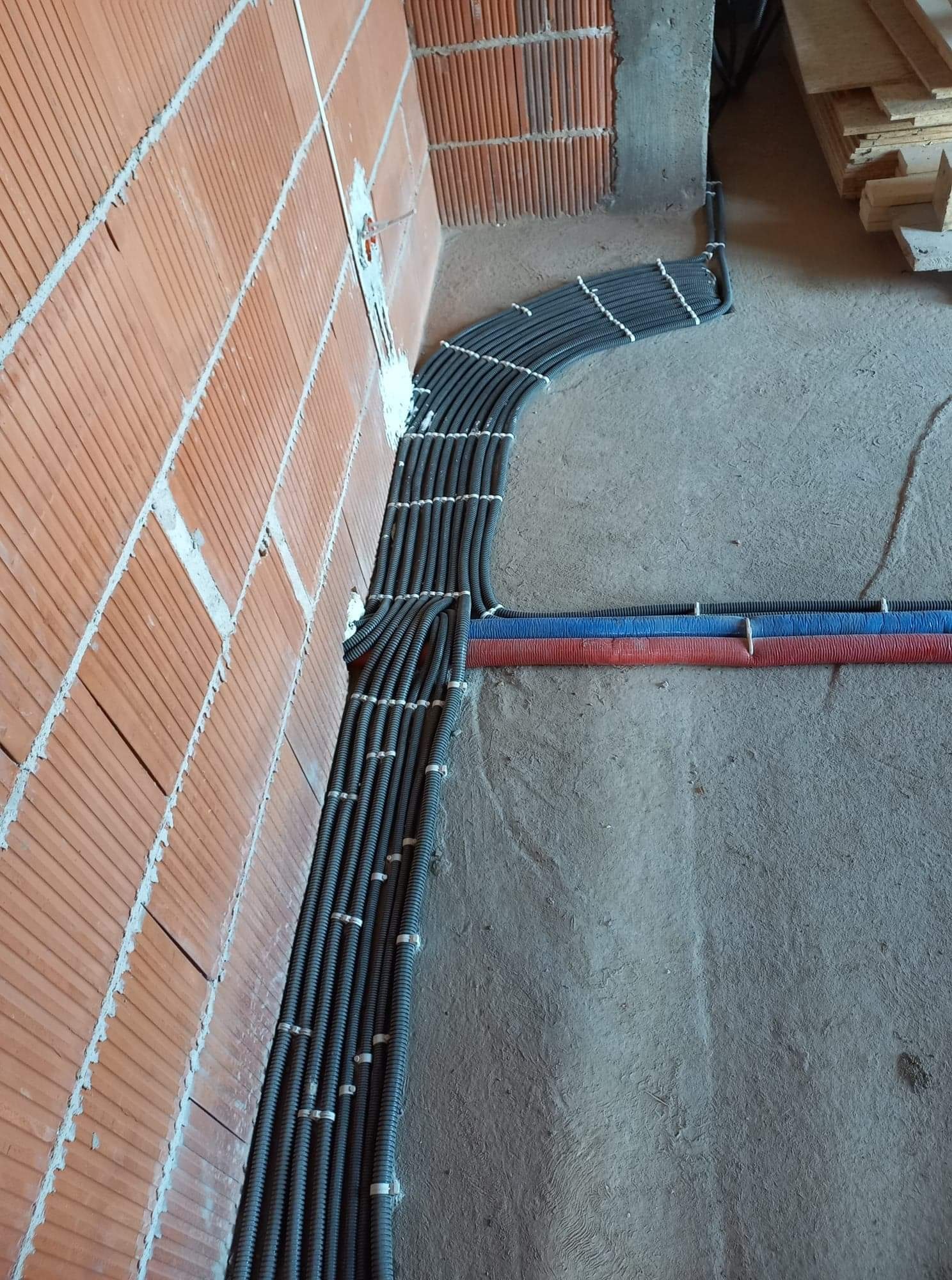 Równolegle ułożone, czarne rury karbowane zabezpieczone białymi uchwytami, prowadzone wzdłuż ceglanej ściany na surowym betonie, z widocznymi fragmentami rur w kolorze czerwonym i niebieskim.