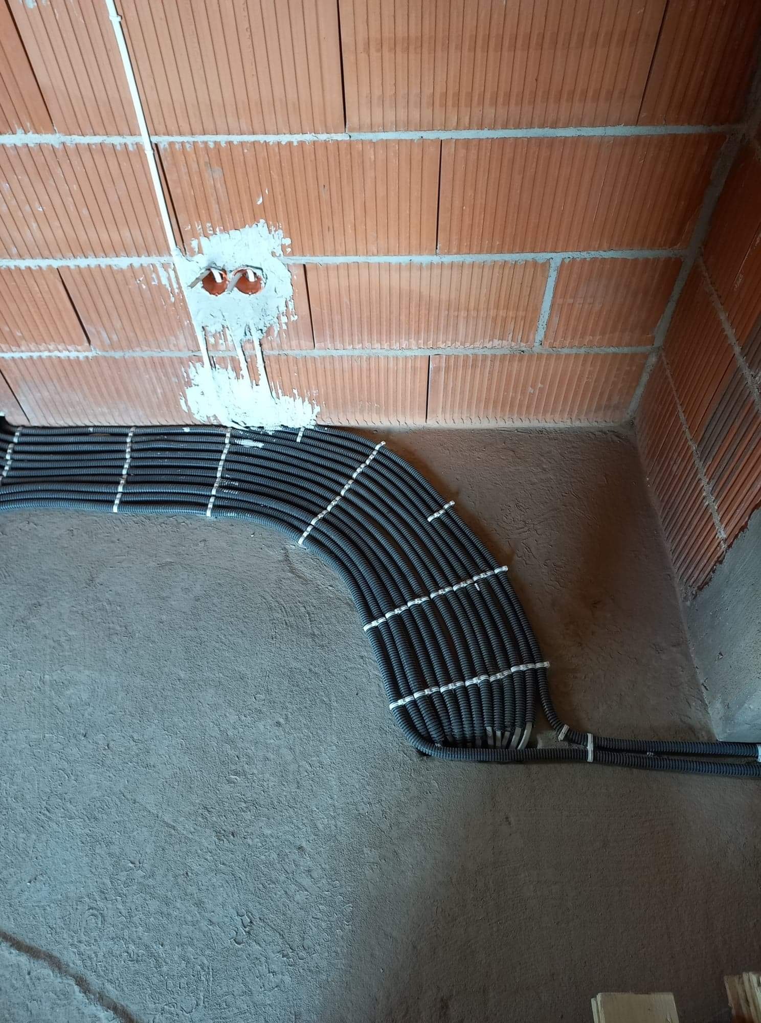 Równolegle ułożone, czarne, karbowane rury instalacyjne, przymocowane białymi opaskami, prowadzone po surowej, betonowej podłodze wzdłuż ceglanej ściany z widocznymi otworami pod gniazdka...