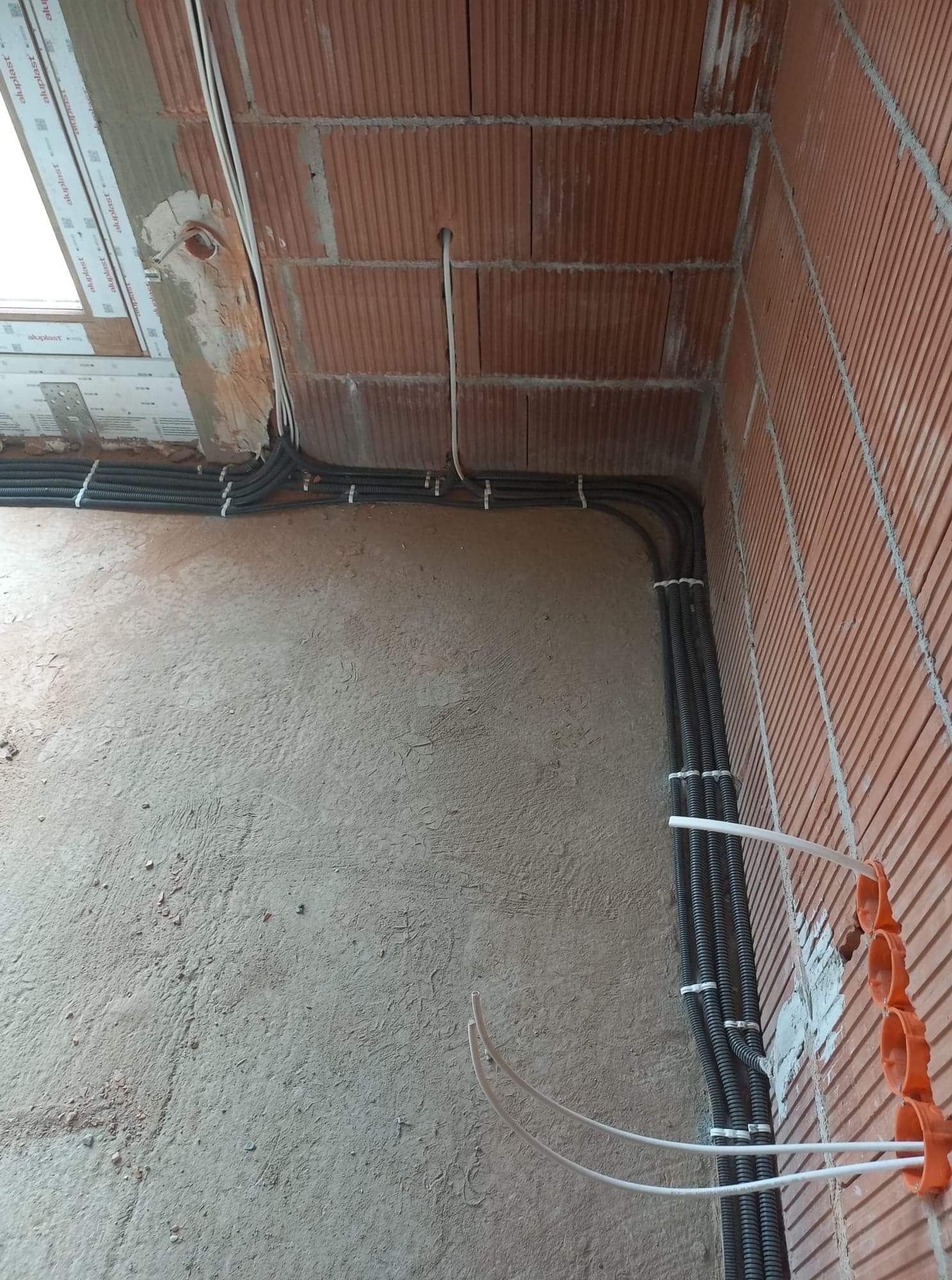 Instalacja elektryczna w nowo budowanym domu: peszle z kablami poprowadzone po ścianach z cegły, widoczne puszki instalacyjne i fragment okna.