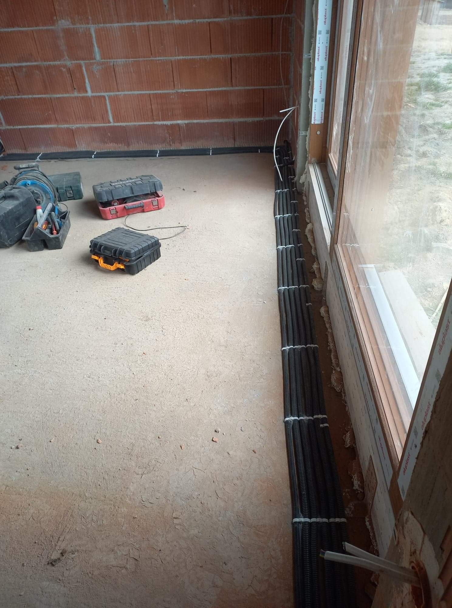 Rzędy czarnych rur karbowanych, zabezpieczonych białymi opaskami zaciskowymi, ułożone wzdłuż okna w surowym stanie budynku z cegły. Widoczne skrzynki narzędziowe w tle.