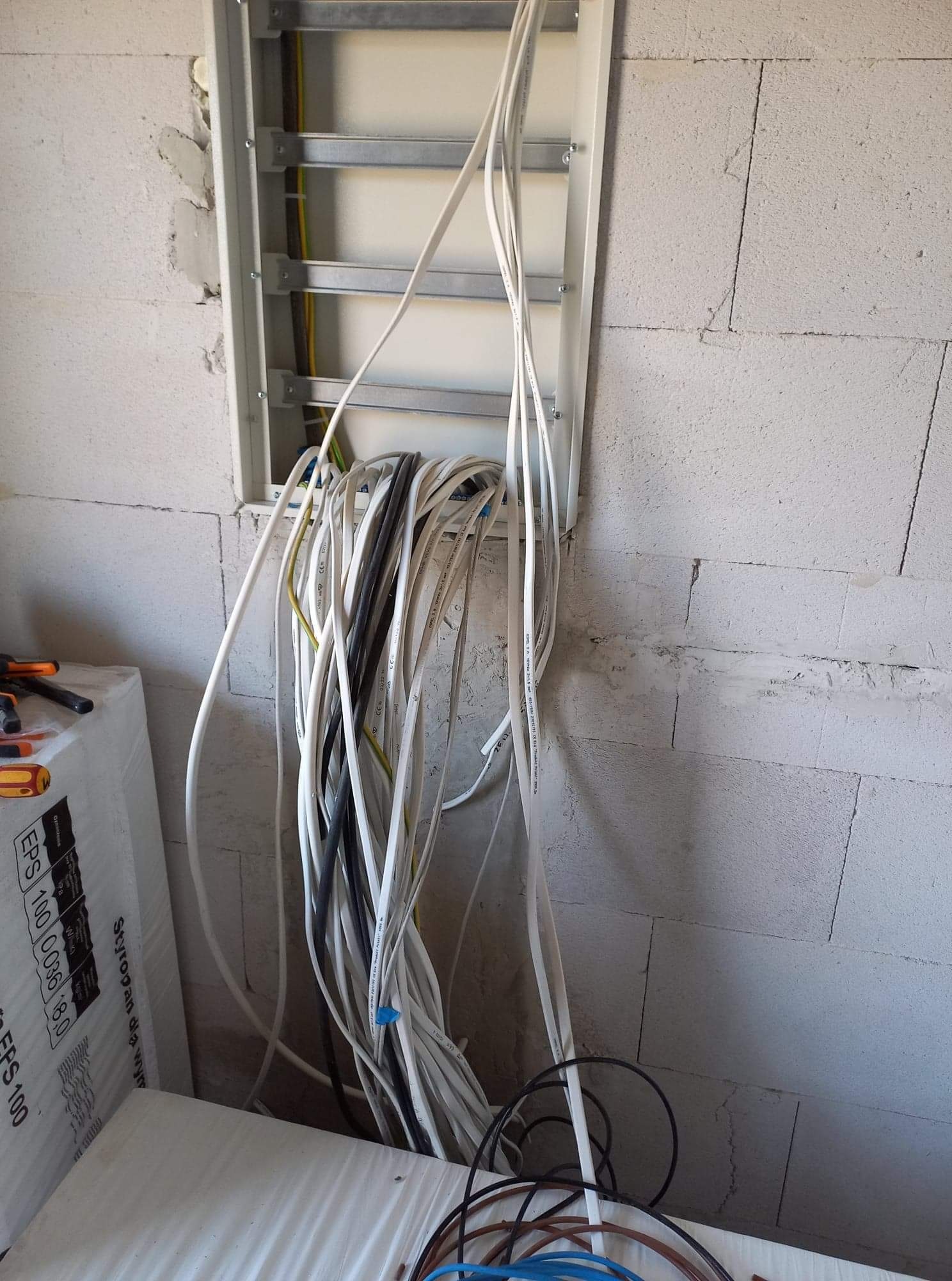 Wiązka kabli elektrycznych wystających z otworu w ścianie z betonu komórkowego, widoczna puszka instalacyjna, obok leżą narzędzia i styropian.