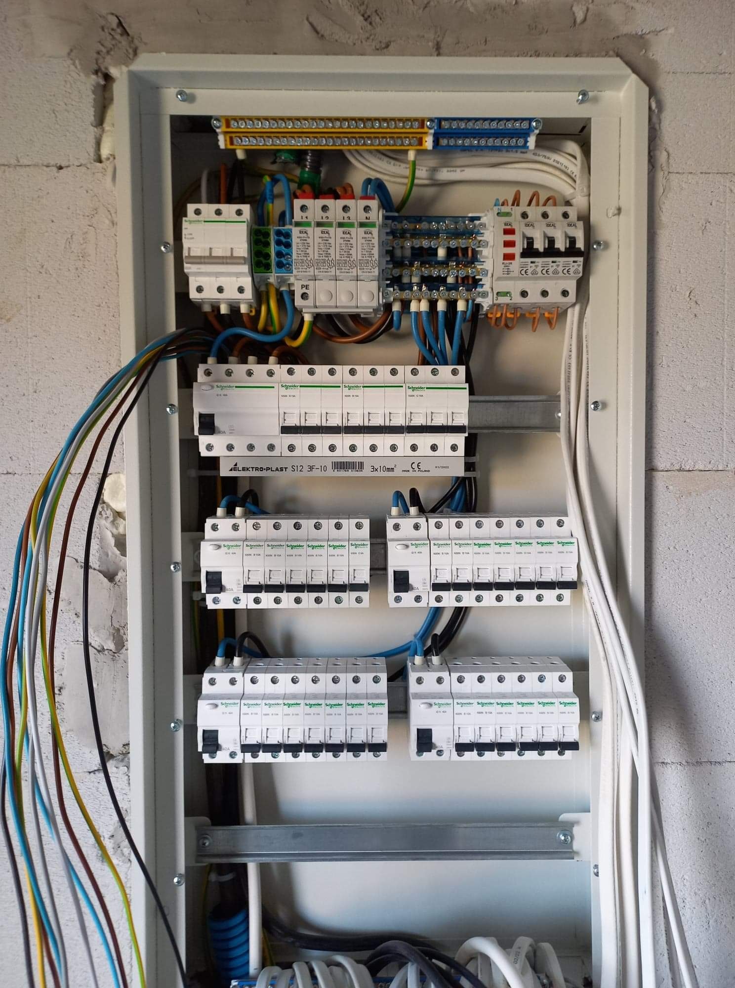 Otwarta skrzynka elektryczna z zamontowanymi bezpiecznikami, przewodami różnej grubości i koloru, listwami zaciskowymi, umieszczona na ścianie z betonu komórkowego.