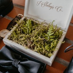Mateusz Gwara Fotografia - Złote obrączki ślubne w białym drewnianym pudełku wypełnionym mchem i paprocią, obok czarne lakierki, mucha i zegarek na brązowym skórzanym pasku.