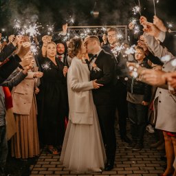 Mateusz Gwara Fotografia - Pocałunek nowożeńców otoczonych gośćmi z zimnymi ogniami na tle wieczornego nieba.