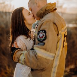 Mateusz Gwara Fotografia - Zakochana para: kobieta w koronkowej sukience i mężczyzna w stroju strażaka, pocałunek na tle zachodzącego słońca.