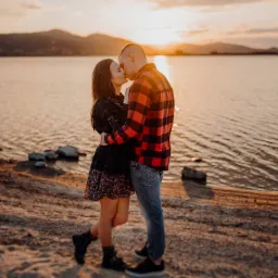 Para całująca się na brzegu jeziora o zachodzie słońca, kobieta w kwiecistej sukience i czarnej kurtce, mężczyzna w czerwonej koszuli w kratę.