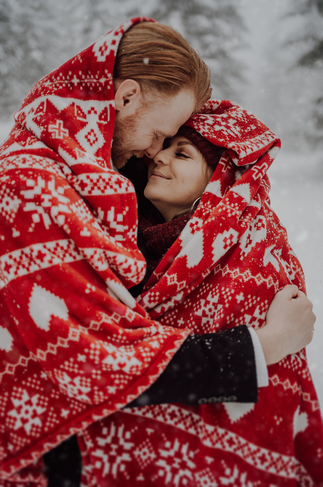 Zimowe ujęcie zakochanej pary otulonej czerwoną, wzorzystą narzutą na tle ośnieżonego krajobrazu.