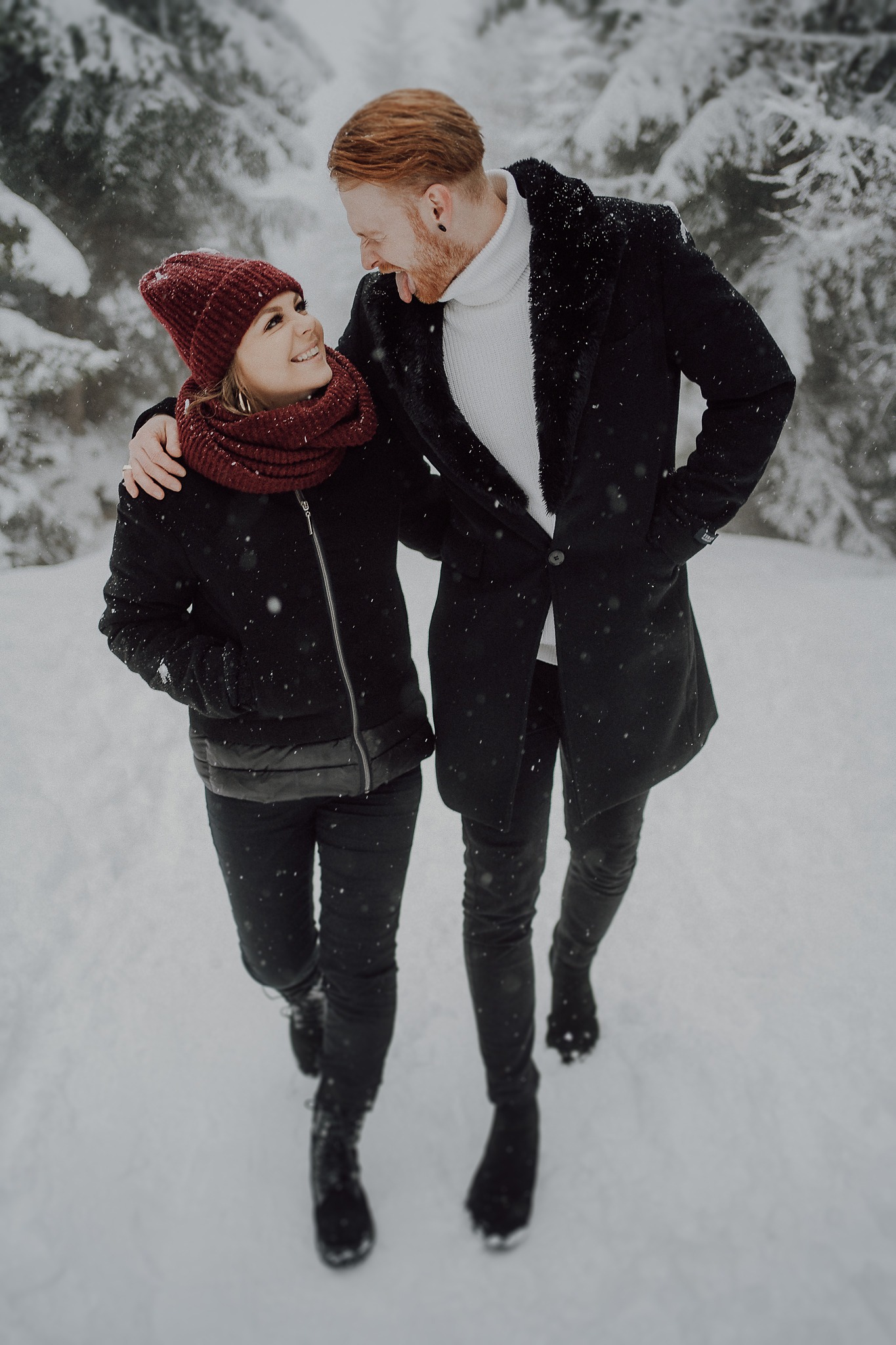 Para zakochanych, kobieta w czerwonej czapce i szaliku, mężczyzna w czarnym płaszczu i białym golfie, spacerują po zasypanym śniegiem lesie, mężczyzna z wyciągniętym językiem patrzy na kobietę.