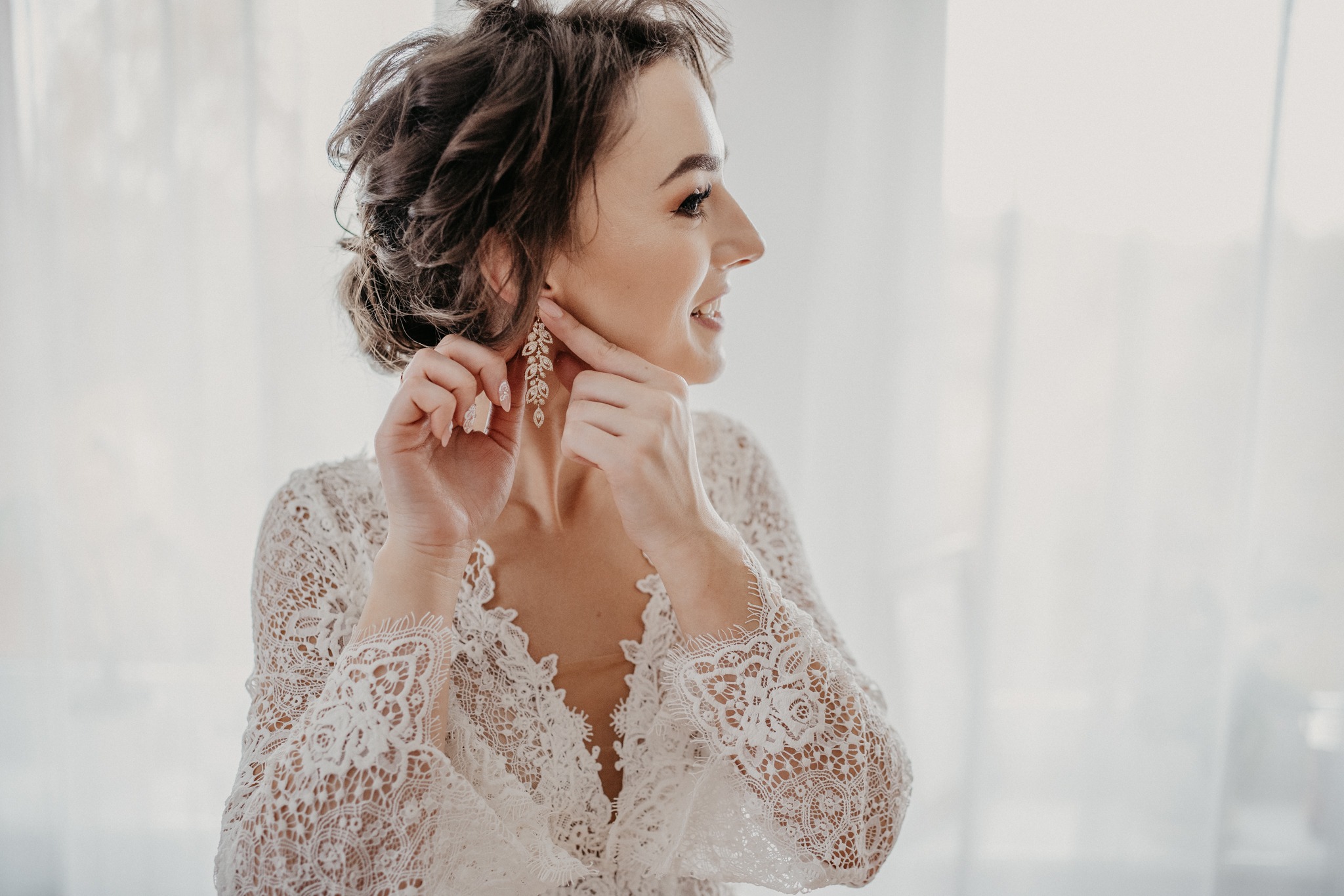 Panna młoda w koronkowej szacie zakłada wiszące, zdobione kolczyki, uśmiechając się w jasnym świetle okna.