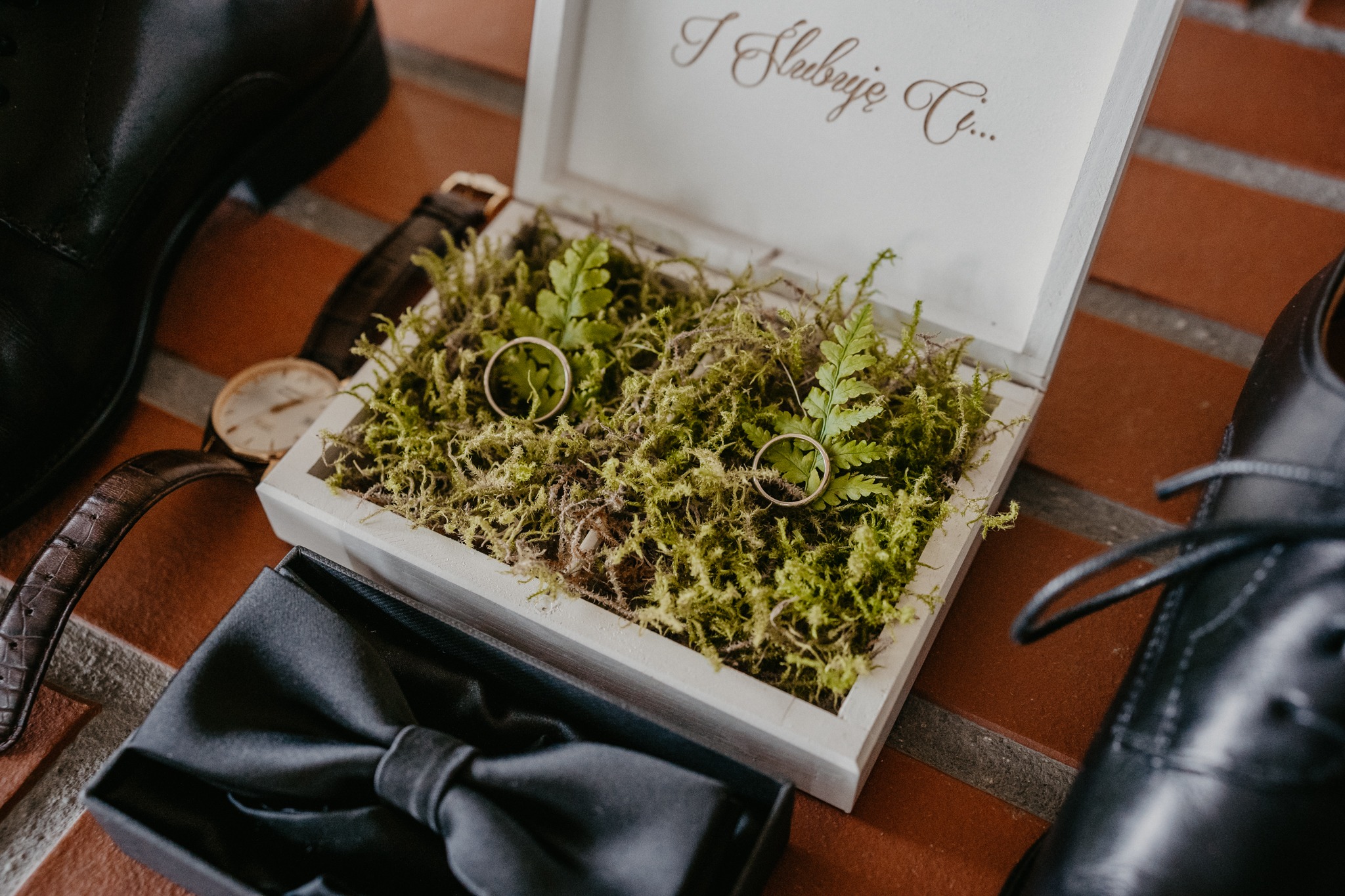 Złote obrączki ślubne w białym drewnianym pudełku wypełnionym mchem i paprocią, obok czarne lakierki, mucha i zegarek na brązowym skórzanym pasku.