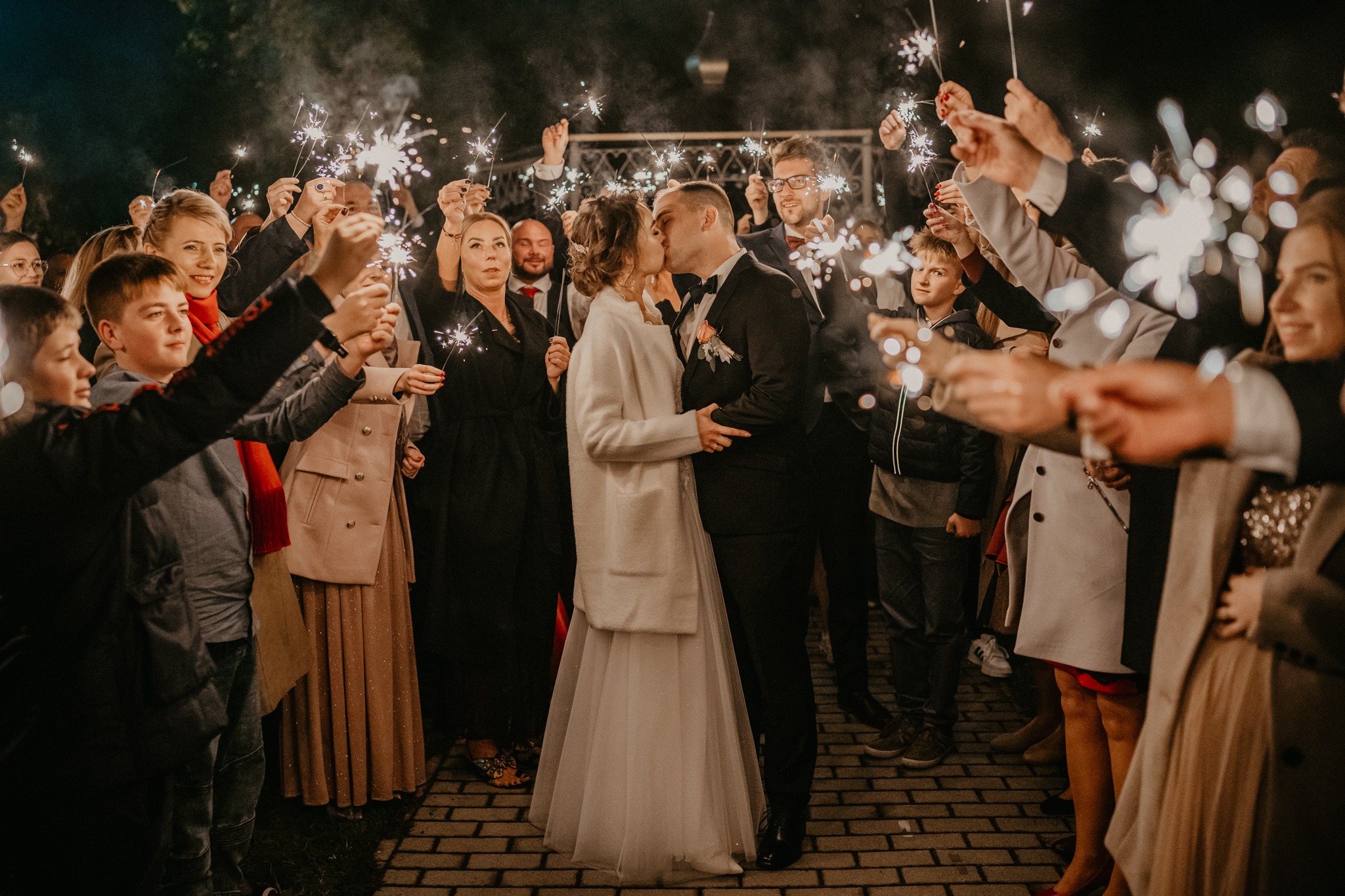 Pocałunek nowożeńców otoczonych gośćmi z zimnymi ogniami na tle wieczornego nieba.