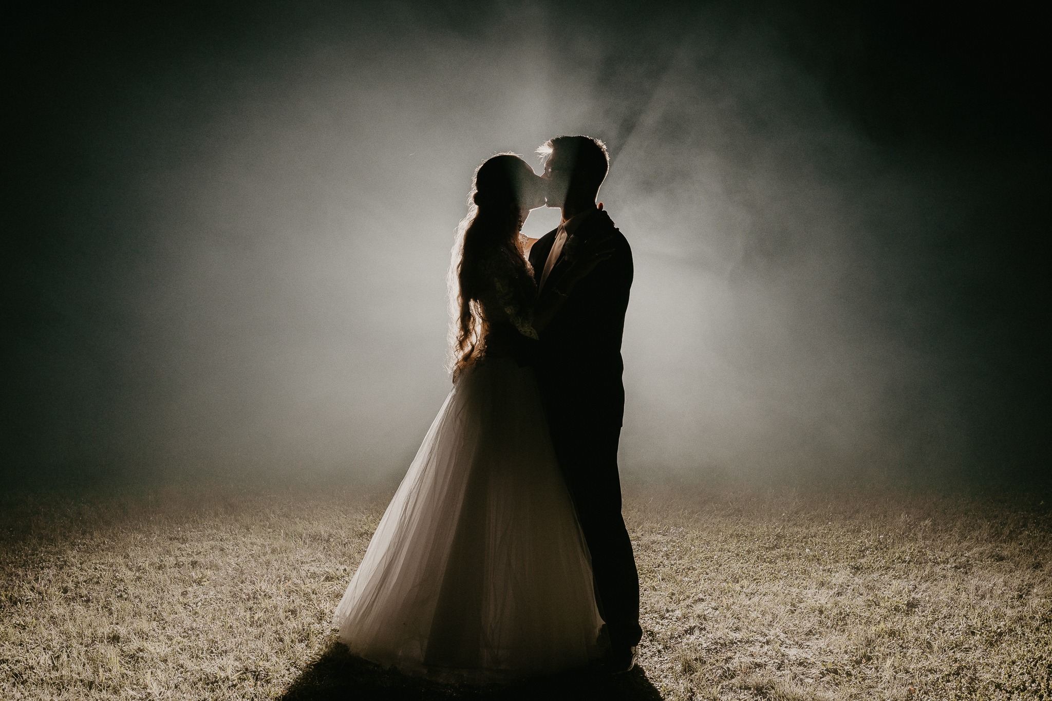 Silhouettes of a bride and groom kissing in a misty, atmospheric wedding photo with a dramatic spotlight effect.