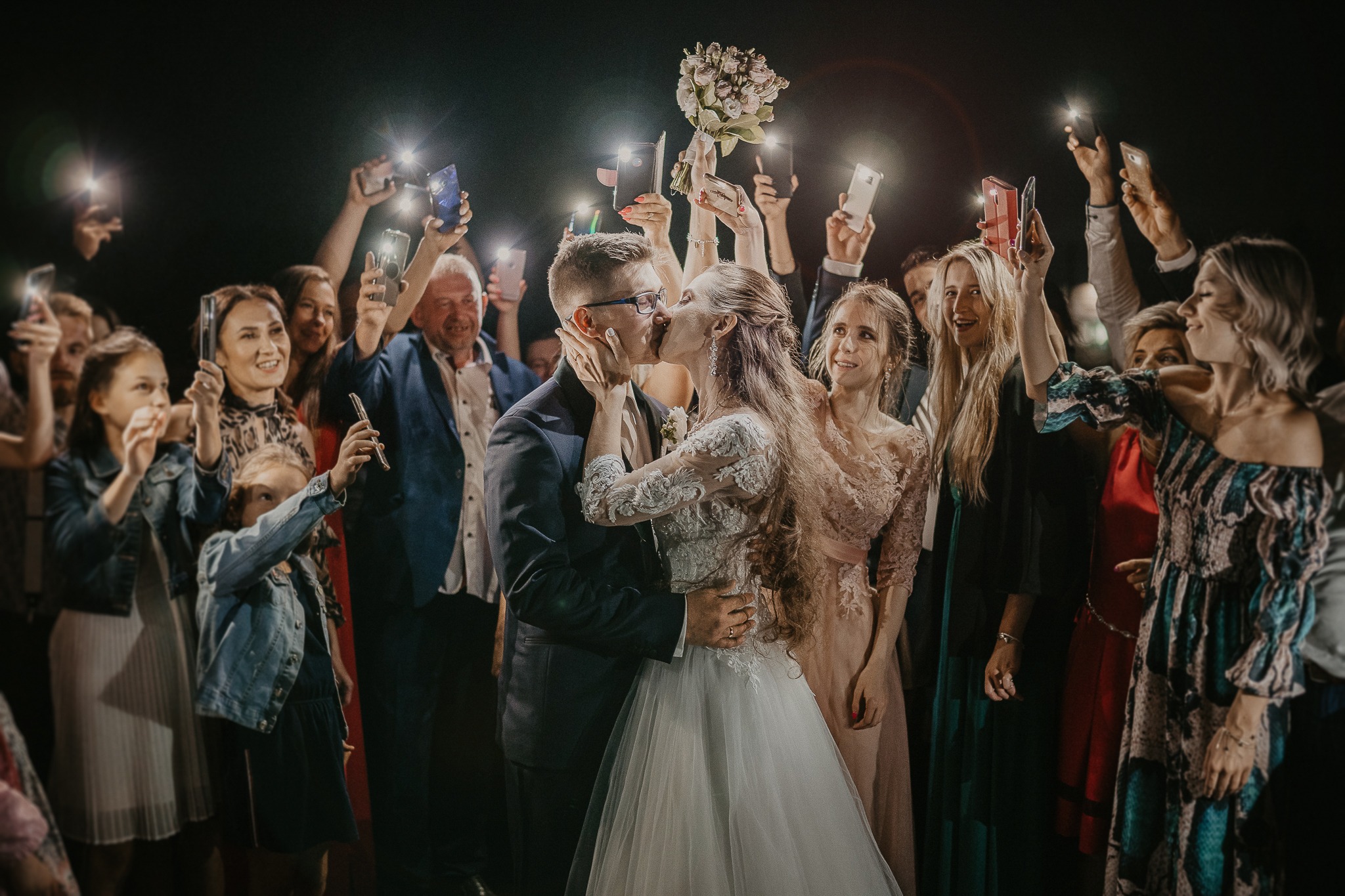 Pocałunek nowożeńców otoczonych gośćmi weselnymi trzymającymi telefony z włączonymi lampami błyskowymi, panna młoda w koronkowej sukni ślubnej, pan młody w ciemnym garniturze.