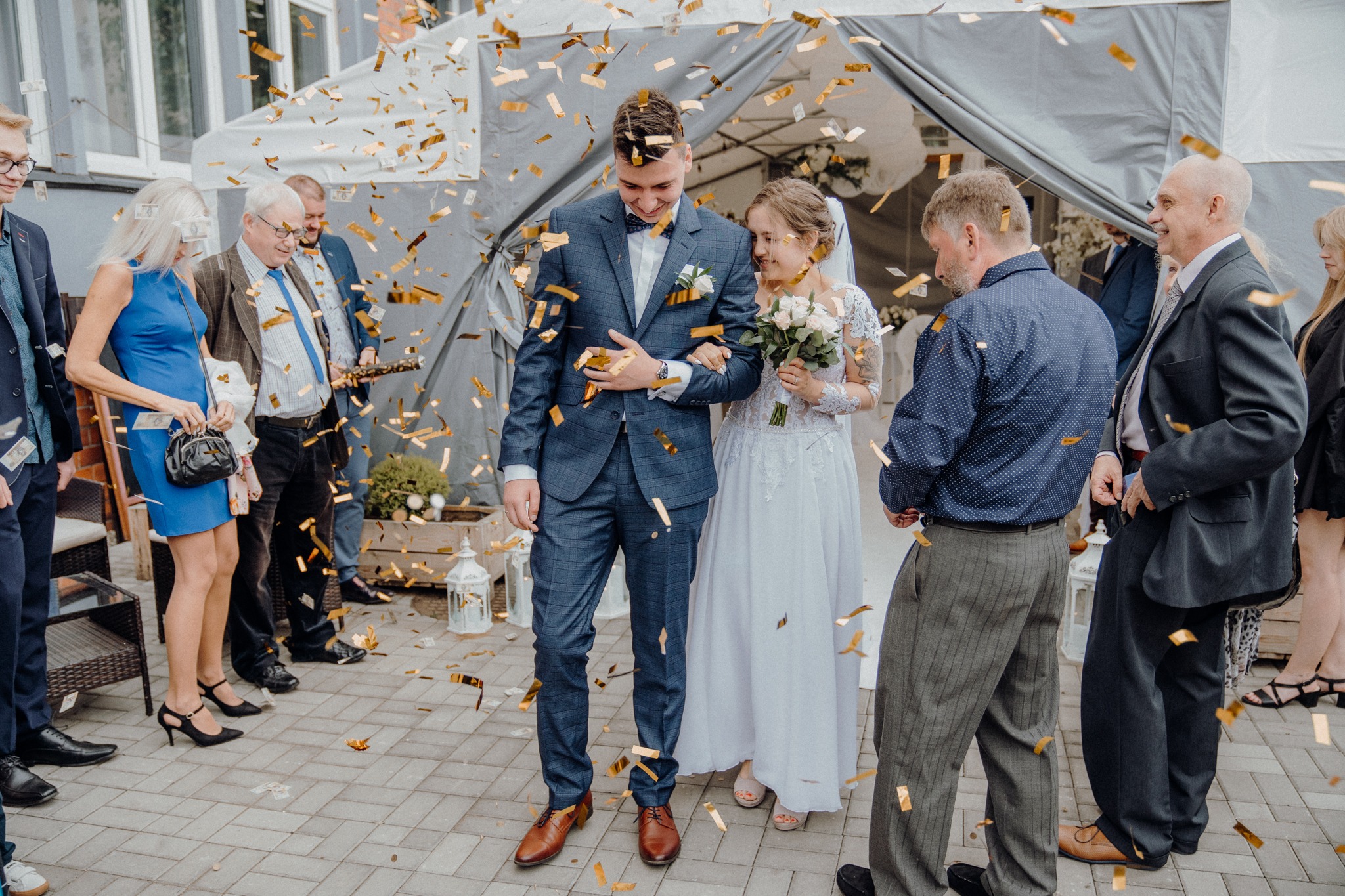 Para młoda w Jeleniej Górze wychodzi spod baldachimu, obsypana złotym konfetti, uśmiechnięta i trzymająca się za ręce, otoczona gośćmi weselnymi.