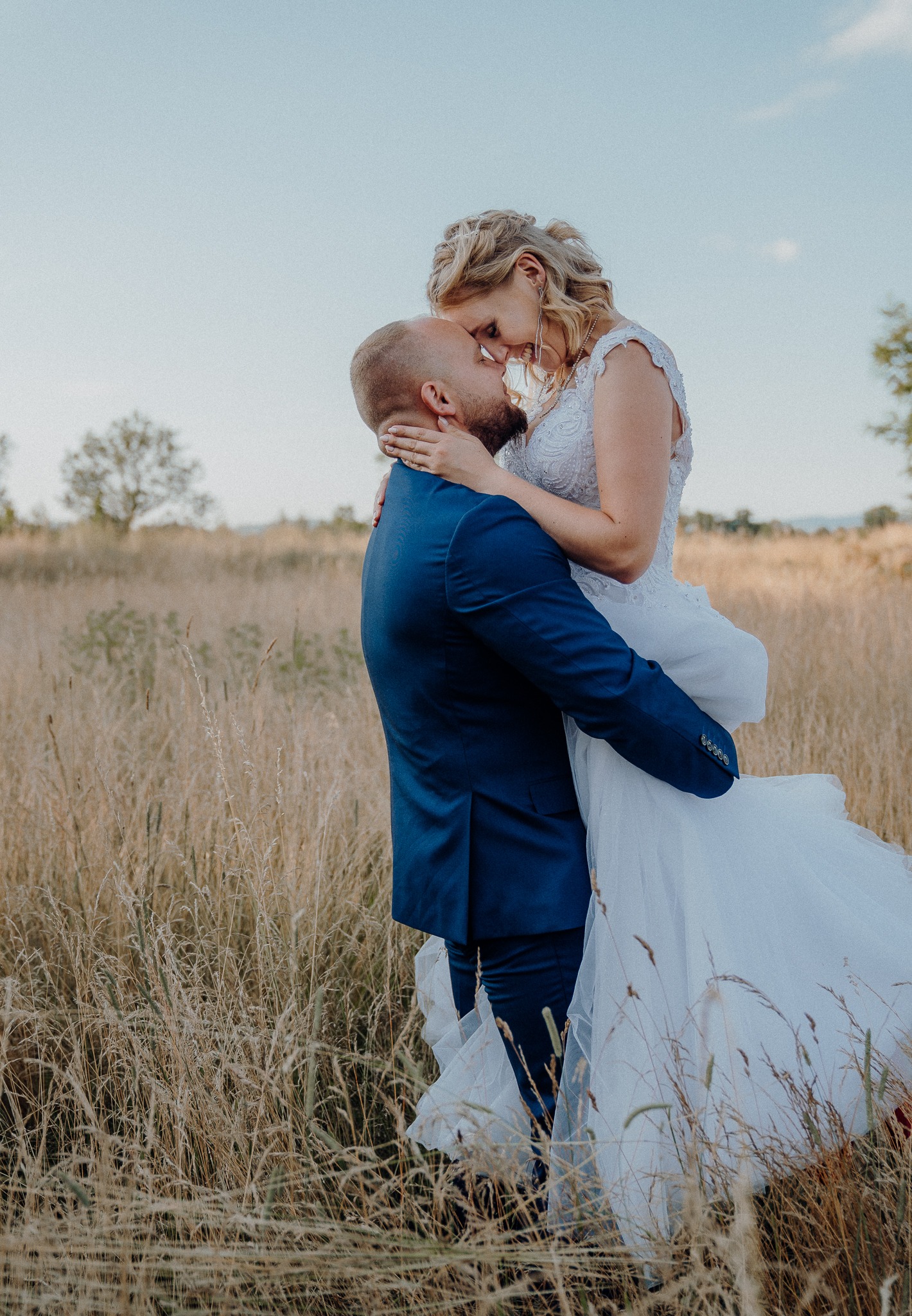 Panna młoda w koronkowej sukni i pan młody w granatowym garniturze, obejmujący się na tle pola z wysoką trawą podczas sesji ślubnej.