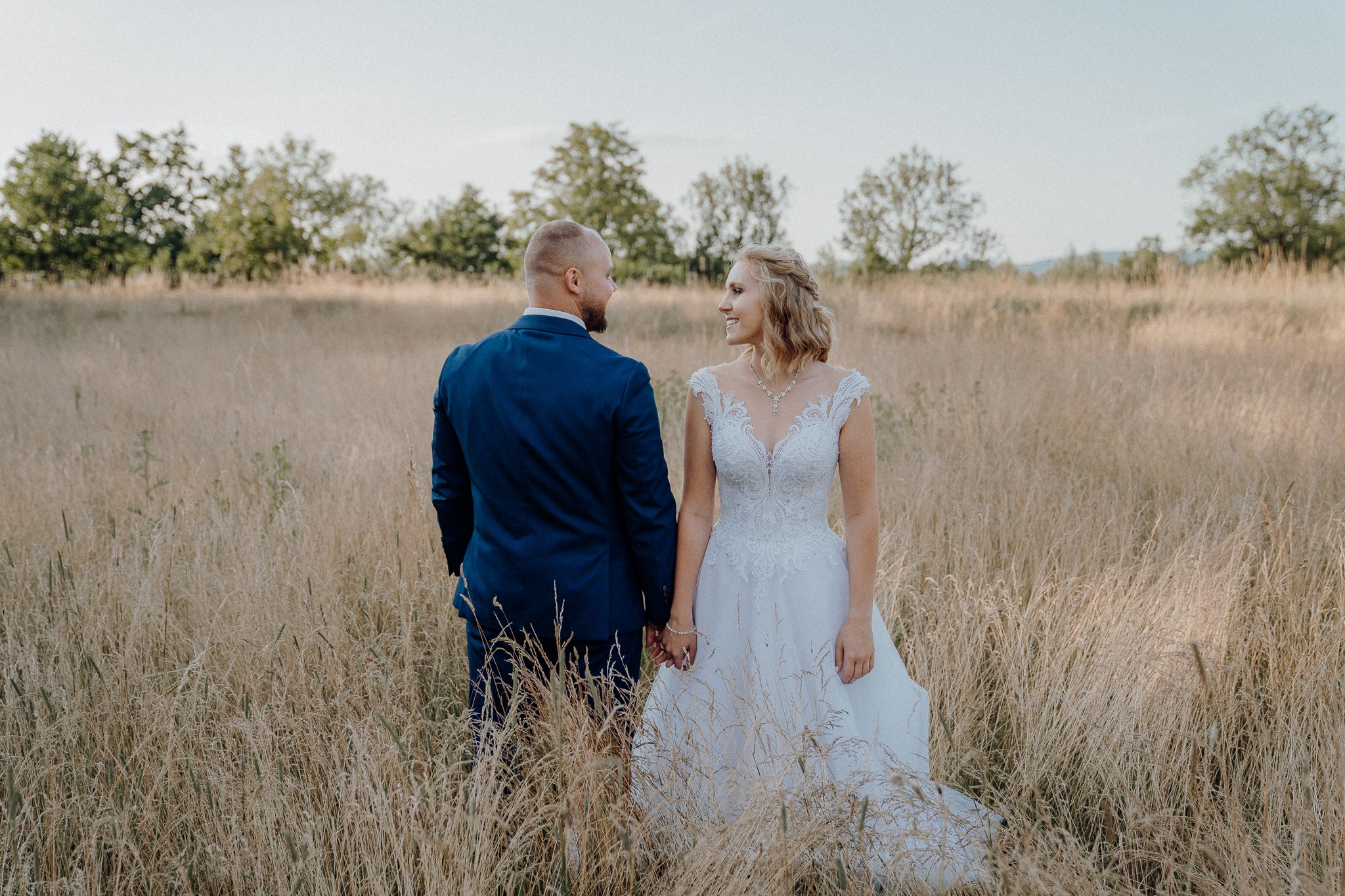 Para młoda trzymająca się za ręce, stojąca na polu z wysoką trawą, on w granatowym garniturze, ona w białej sukni ślubnej z koronkowym dekoltem, oboje patrzą na siebie.